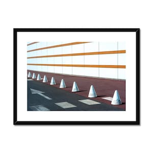 Könnte beinhalten: Gerahmtes Foto einer modernen Architekturszene. Weiße Verkehrskegel säumen eine dunkle Asphaltstraße mit einem weißen Pfeil, der nach links zeigt. Ein roter Gehweg grenzt an ein Gebäude mit weißen und orangefarbenen horizontalen Streifen.