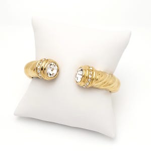May include: A gold-toned bangle bracelet with two clear crystal stones set in the center of each end. The bracelet has a twisted design.