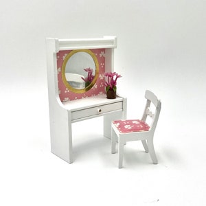 May include: Miniature white vanity set with a round gold-framed mirror, a drawer, and a matching chair. The vanity has a pink floral patterned backing and a small pot of pink flowers. The chair has a pink floral patterned seat.