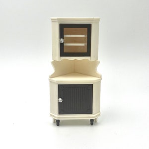 May include: A miniature corner cabinet in cream and dark brown. The upper section has a glass-fronted door with two shelves, while the lower section features a solid door with decorative detailing. The cabinet has small black feet.