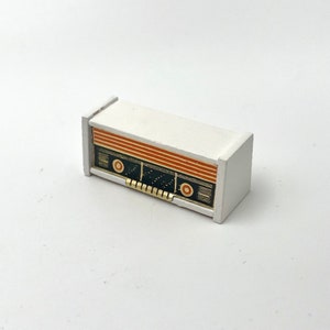 May include: A miniature white radio with orange and black detailing. The radio has a rectangular shape with a speaker grill and control knobs. The design is reminiscent of vintage radios.