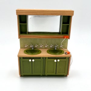 May include: A green bathroom vanity with two sinks, a mirror, and a wooden cabinet. The vanity has two white knobs on each door and a white tile backsplash with floral designs.