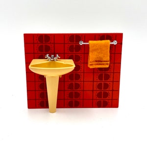 May include: A miniature bathroom set with a yellow sink and pedestal, a towel rack, and a brown and orange tiled wall.