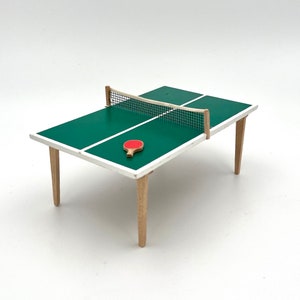 Op de afbeelding: Kleine pingpongtafel met een groen oppervlak, witte lijnen en een witte rand. Het heeft houten poten en een zwart net. Een rode tafeltennisbat ligt op de tafel. Het is een speelgoedartikel.