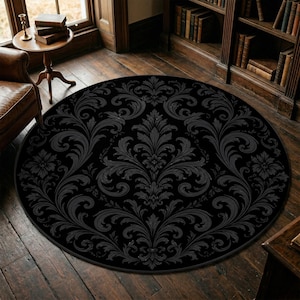 Puede incluir: Una alfombra redonda negra con un diseño floral damasco gris. La alfombra está centrada en un suelo de madera oscura, con un sillón de cuero y una pequeña mesa con libros en el fondo. El diseño de la alfombra es simétrico.