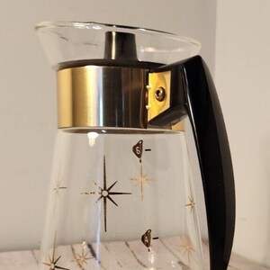 May include: A glass coffee pot with a gold band around the top and a black handle. The pot has a starburst design and markings for 2, 4, and 6 cups. The text "CONT. HEAT PROOF GLASS" is printed on the bottom of the pot.