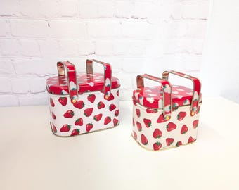 May include: Two red and white metal tins with strawberry patterns and gold handles. The tins are different sizes and are sitting on a white surface.