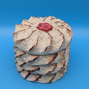 Peut inclure: Une pile de quatre objets ronds décoratifs blanc cassé avec une surface texturée et des bords marron. Un petit objet rouge texturé est posé dessus. Le fond est bleu clair uni.