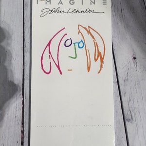 May include: A white cassette tape case with a colourful cartoon drawing of John Lennon's face. The text "Imagine John Lennon" is printed on the front of the case. The price tag reads "£20.99".