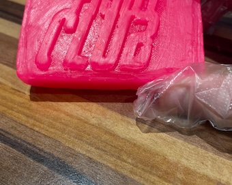 May include: A rectangular bar of bright pink soap with the word "CLUB" embossed on the surface. The soap is held by a gloved hand, and rests on a wooden surface.