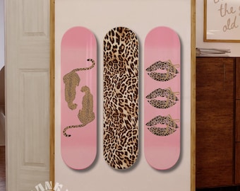 May include: A framed artwork featuring three skateboard decks. The decks have pink and leopard print designs. One deck has two leopards, another has a leopard print pattern, and the third has three sets of lips.