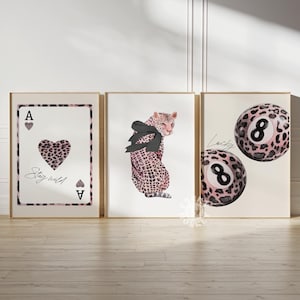 May include: Three framed prints featuring a pink and black leopard print design. The first print shows an ace of hearts playing card with the text "Stay wild". The second print depicts a leopard wearing a black bow. The third print displays two pink and black 8-ball pool balls with the text "Lucky".