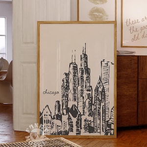 Può includere: Una stampa artistica incorniciata con un'illustrazione in bianco e nero dello skyline di Chicago. La parola "chicago" è scritta in un carattere corsivo sotto gli edifici. La stampa è in una cornice di legno chiaro. Altre due stampe incorniciate sono visibili sullo sfondo.