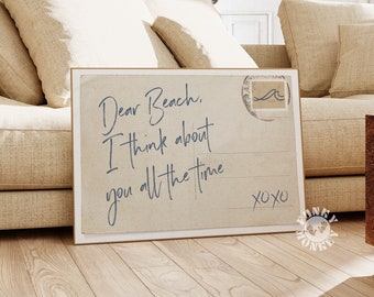 May include: A vintage-style postcard with the text "Dear Beach, I think about you all the time XOXO" printed on it. The postcard is framed and leaning against a beige couch.