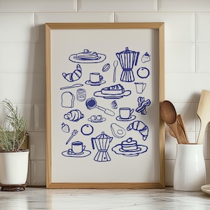 May include: A framed print featuring a blue line drawing of breakfast items, including coffee cups, croissants, pancakes, and a coffee pot. The artwork is set against a cream background and framed in light wood. The print is displayed on a white tiled surface.