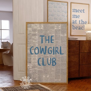 May include: A framed print with a newspaper background and the text "THE COWGIRL CLUB" in blue lettering. The print is hanging on a wall in a room with a wooden floor and a wooden dresser.