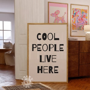 Op de afbeelding: Een ingelijst print met een witte achtergrond en zwarte tekst die "COOL PEOPLE LIVE HERE" luidt. De print hangt aan een muur in een kamer met een houten vloer en een houten kast.
