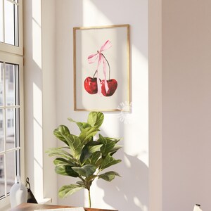 May include: A watercolor painting of two red cherries tied together with a pink ribbon. The cherries are hanging from a branch and the ribbon is tied in a bow. The painting is framed in a light wood frame.