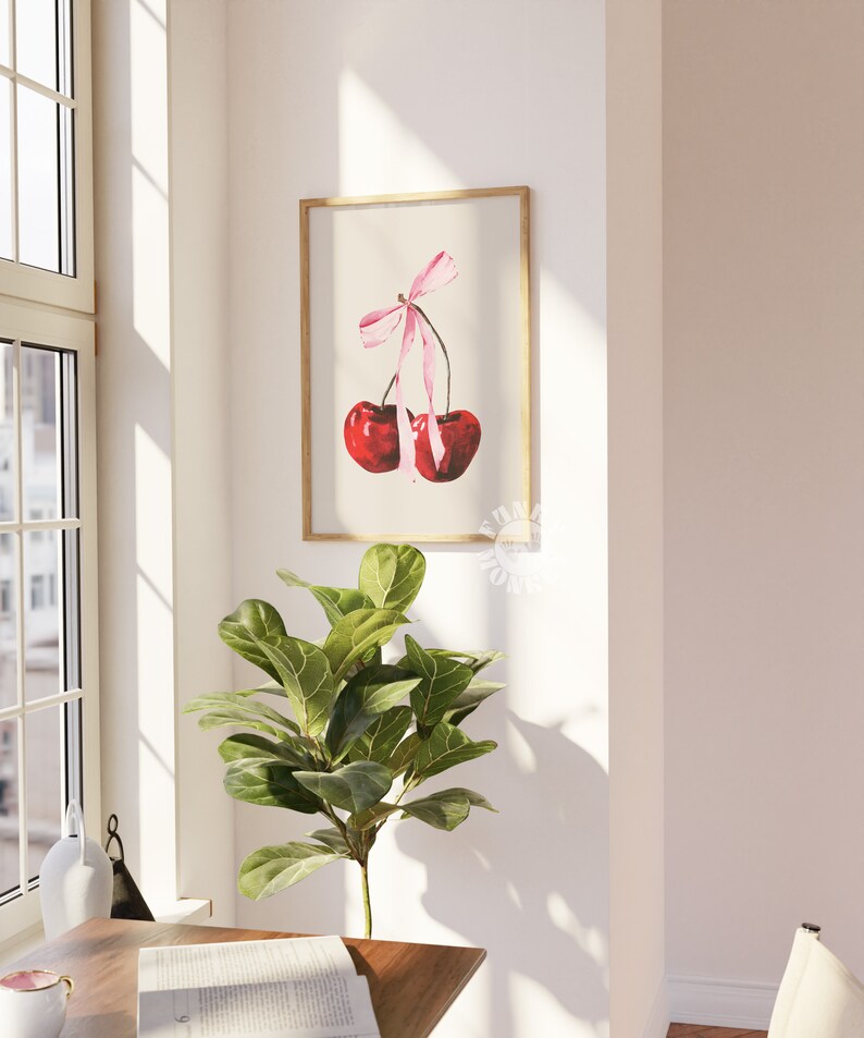 May include: A watercolor painting of two red cherries tied together with a pink ribbon. The cherries are hanging from a branch and the ribbon is tied in a bow. The painting is framed in a light wood frame.