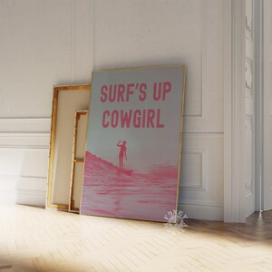 May include: A pink and white poster with the text "SURF'S UP COWGIRL" in pink letters. The poster features a silhouette of a person surfing on a pink wave.