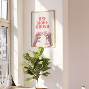 Peut inclure: Une impression encadr&eacute;e avec le texte "ONE MORE ROUND" en lettres roses. Deux mains tiennent des verres de vin, l'un rouge et l'autre blanc, sur un fond blanc.
