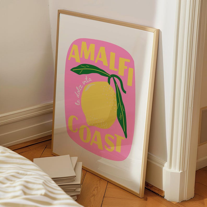 May include: A framed art print featuring a yellow lemon with green leaves on a pink background. The words "AMALFI COAST" are in yellow, with "la dolce vita" in cursive. The print is in a gold frame.