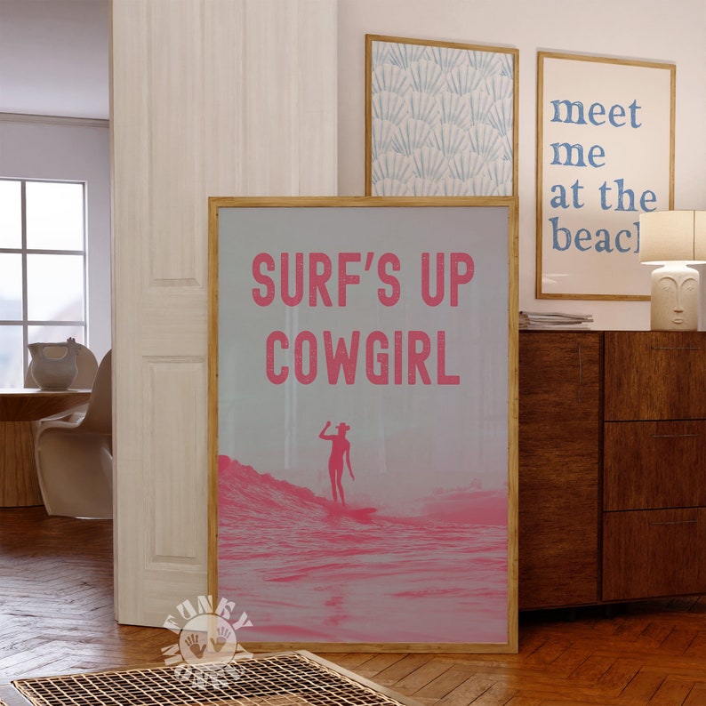 May include: A pink and white poster with the text "Surf's Up Cowgirl" in pink letters. A silhouette of a person wearing a cowboy hat and holding a surfboard is surfing on a pink wave.