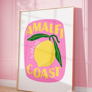 May include: A framed art print featuring a yellow lemon with green leaves against a pink background. The words "AMALFI COAST" are in yellow, with "la dolce vita" in cursive. The print is in a white frame.