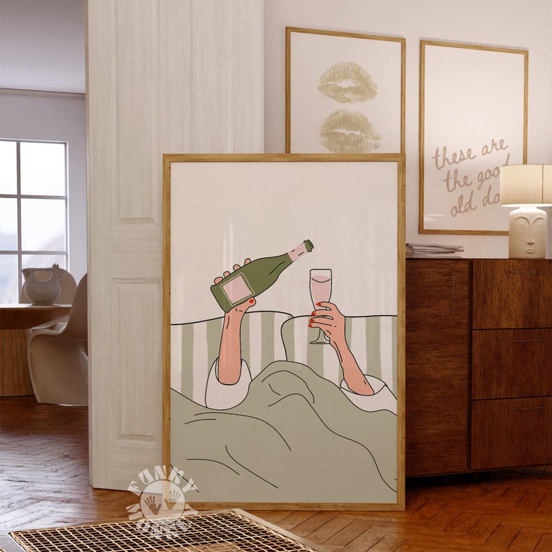 May include: A framed art print depicts a person pouring champagne into a glass while in bed. The illustration features a green champagne bottle, a pink drink, and a green and white striped background. Additional framed art pieces are visible.