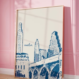 May include: A framed art print featuring a blue and white illustration of the Austin, Texas skyline. The word "austin" is written in cursive. The artwork is displayed against a cream-colored background and framed in a light wood.