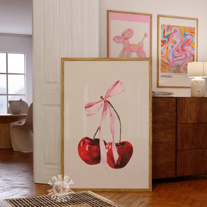 May include: A framed print of two red cherries with a pink ribbon bow. The cherries are painted in a watercolor style and the background is a light cream color.