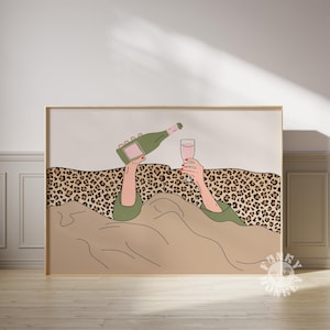 K&ouml;nnte beinhalten: Ein gerahmter Kunstdruck zeigt eine Person, die Champagner in ein Glas gie&szlig;t. Die Illustration zeigt eine gr&uuml;ne Champagnerflasche, ein Glas rosa Fl&uuml;ssigkeit und H&auml;nde mit rotem Nagellack. Der Hintergrund umfasst einen Leopardenmuster und eine beige Decke.