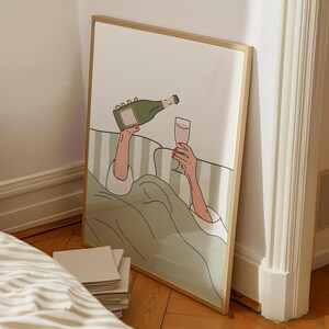 May include: A framed art print depicts a person pouring champagne into a glass while in bed. The illustration features a green champagne bottle, a glass of pink liquid, and a green and white striped background.