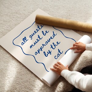 May include: A white poster with blue text that reads "all guests must be approved by the cat". A person is rolling up the poster.
