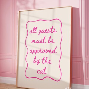 May include: A pink and white framed print with the text "all guests must be approved by the cat" in a pink cursive font.