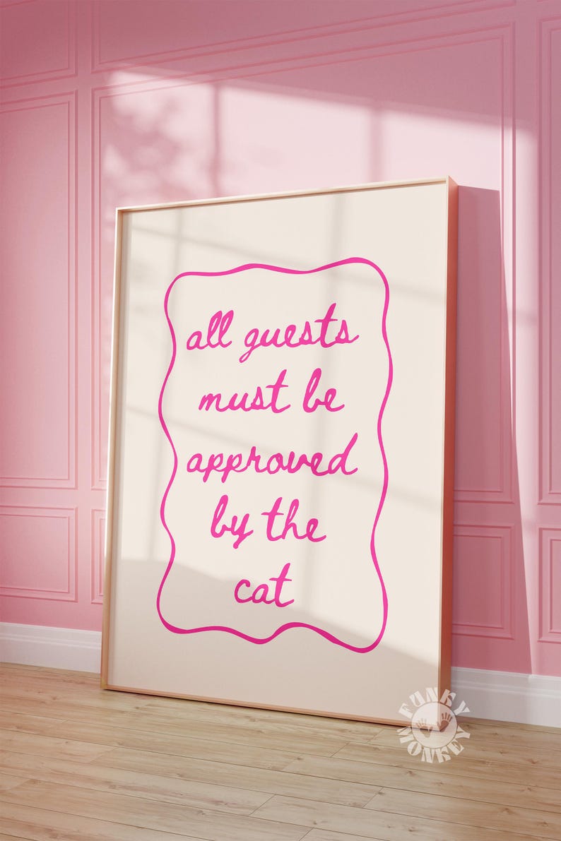 May include: A pink and white framed print with the text "all guests must be approved by the cat" in a pink cursive font.