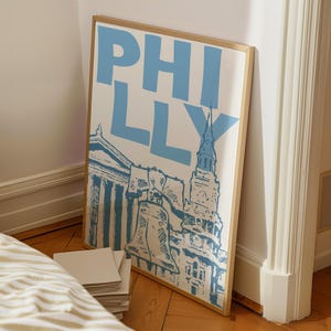 May include: A framed art print featuring the word "PHILLY" in large, light blue letters. Below the text is a detailed illustration of Philadelphia landmarks in blue and white. The artwork is displayed in a room with a wooden floor.