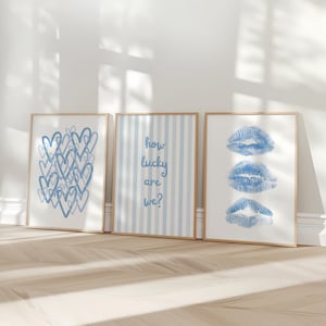 May include: Three framed art prints with a light wood frame. The prints feature blue heart shapes, blue striped background with the text "how lucky are we?", and three blue lip prints. The prints are displayed on a light wood floor.