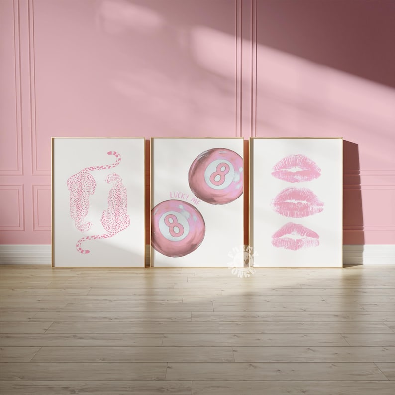 May include: Three framed art prints with gold frames. The prints feature pink leopard illustrations, pink eight-ball designs with the text "LUCKY ME", and pink lip prints. The artwork is displayed against a pink wall and wooden floor.