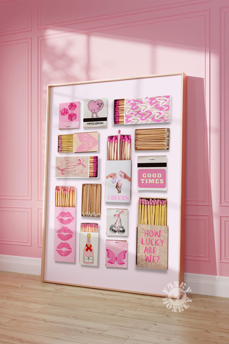 May include: A pink and white framed collage of matchboxes with various designs, including hearts, dice, cherries, a butterfly, lips, and text that reads "Good Times", "Cheers", and "How Lucky Are We?"