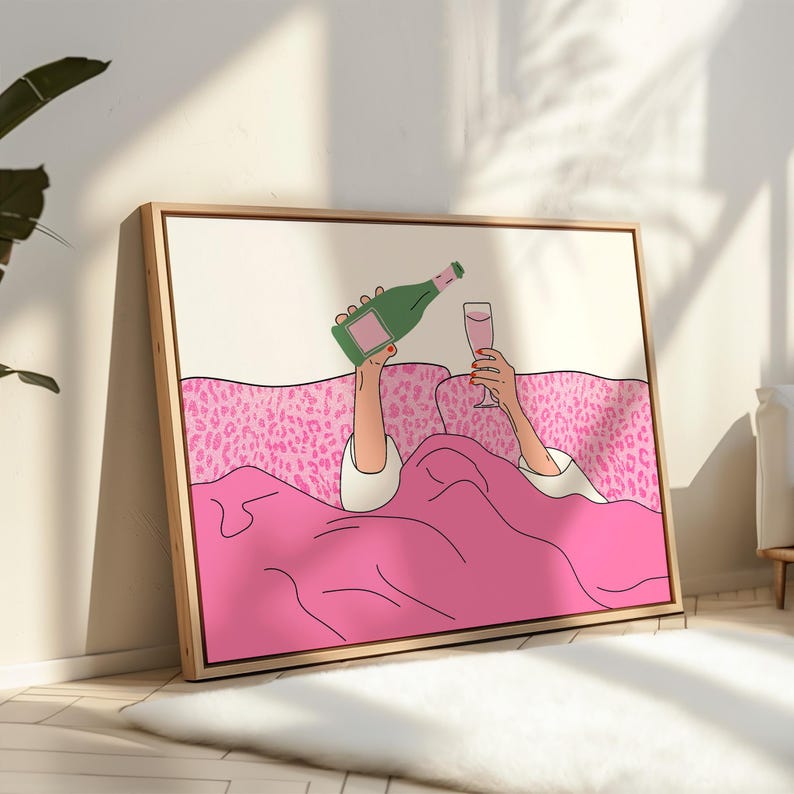 May include: A framed art print depicts a person in bed pouring champagne into a glass. The illustration features a pink blanket, leopard-print pillows, and a green champagne bottle. The artwork has a minimalist style with a light wood frame.