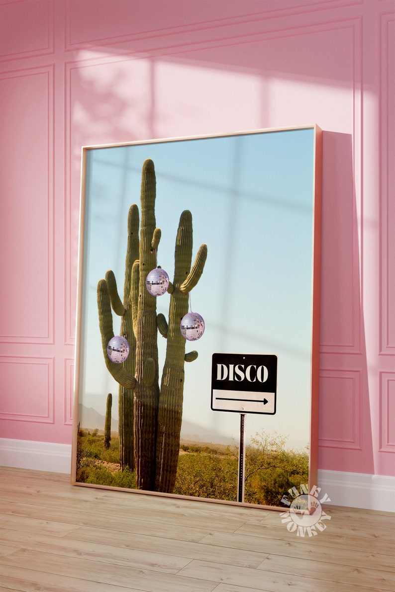 May include: A framed print showing a saguaro cactus decorated with disco balls. A sign reading "DISCO" points right. The background depicts a desert landscape under a clear blue sky. The frame is gold.