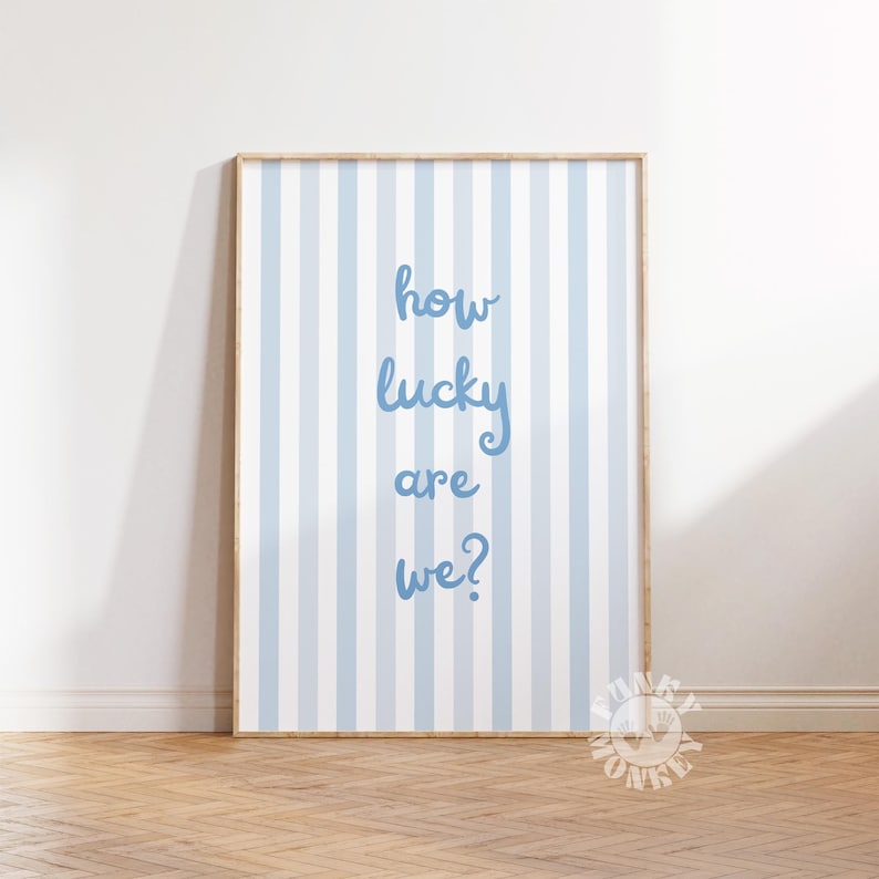 May include: Framed wall art with a light blue and white striped background. The text "how lucky are we?" is written in a cursive font in light blue. The frame is a light wood color.