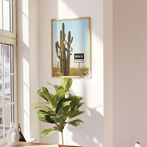 May include: A framed print depicting a desert scene with a large saguaro cactus decorated with disco ball baubles. A signpost indicates "DISCO" against a blue sky. The artwork is displayed in a room with a window and a potted plant.
