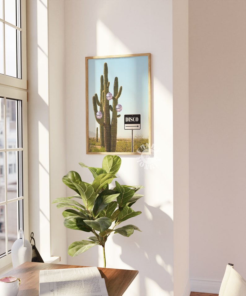 May include: A framed print depicting a desert scene with a large saguaro cactus decorated with disco ball baubles. A signpost indicates "DISCO" against a blue sky. The artwork is displayed in a room with a window and a potted plant.