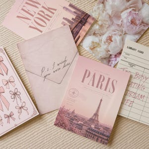 May include: A collection of stationery items including a New York postcard, a floral print, a pink envelope with the text P.S. I wish you were here, a Paris postcard featuring the Eiffel Tower, and a library card with the text Too busy creating dream life.