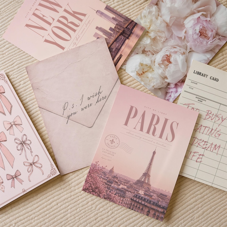 May include: A collection of stationery items including a New York postcard, a floral print, a pink envelope with the text P.S. I wish you were here, a Paris postcard featuring the Eiffel Tower, and a library card with the text Too busy creating dream life.