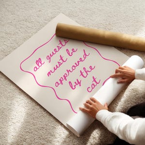 May include: A white poster with pink lettering that reads "All guests must be approved by the cat". A person is rolling up the poster.