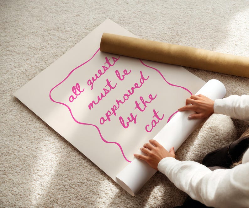 May include: A white poster with pink lettering that reads "All guests must be approved by the cat". A person is rolling up the poster.