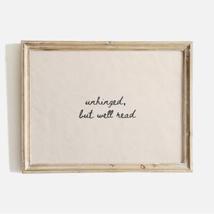 May include: A rectangular wall art piece featuring a light beige background inside a weathered light brown wooden frame. The text "unhinged, but well read" is printed in the center in a black, handwritten-style cursive font.